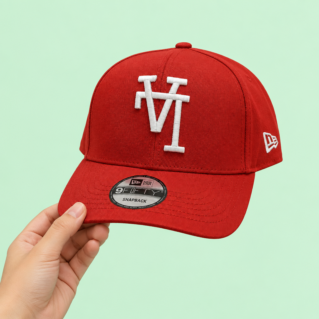 Red cap with white raised embroidery held in one hand, side-forward view on pastel mint background